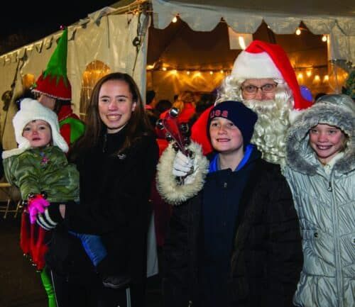 Family and Santa posing together at festive holiday market tent with warm lights.
