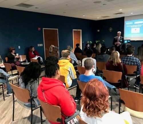 High school students seated in circle listening to addiction recovery presentation by Kettering Health professionals.