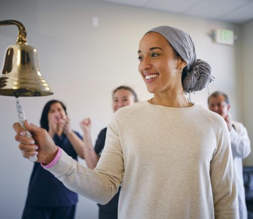 An adult woman chemotherapy patient in a treatment office, celebrating the completion of her treatment with a ceremonial bell ring.