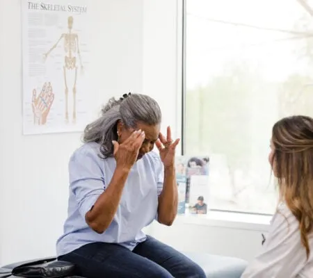 An unrecognizable senior adult woman touches her temples as she explains her headache symptoms to the unrecognizable female doctor.
