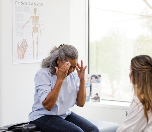 An unrecognizable senior adult woman touches her temples as she explains her headache symptoms to the unrecognizable female doctor.