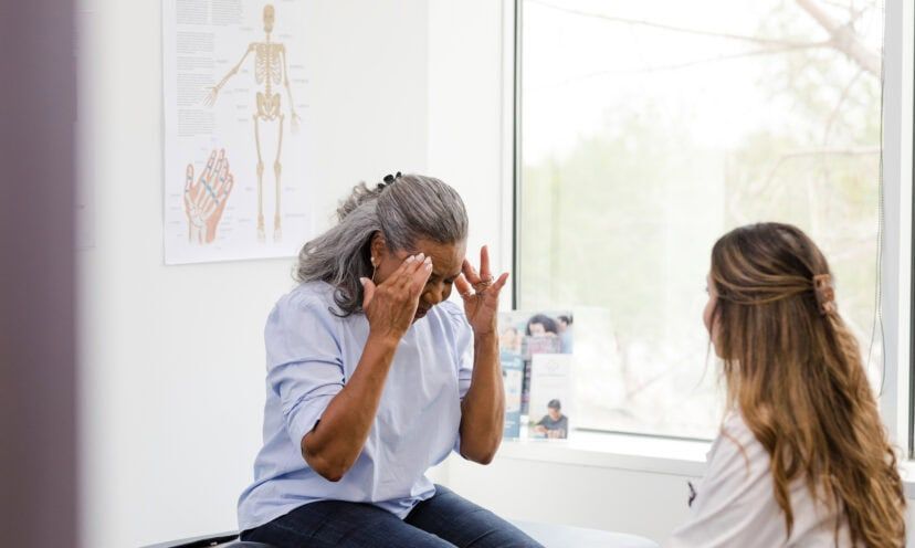 An unrecognizable senior adult woman touches her temples as she explains her headache symptoms to the unrecognizable female doctor.
