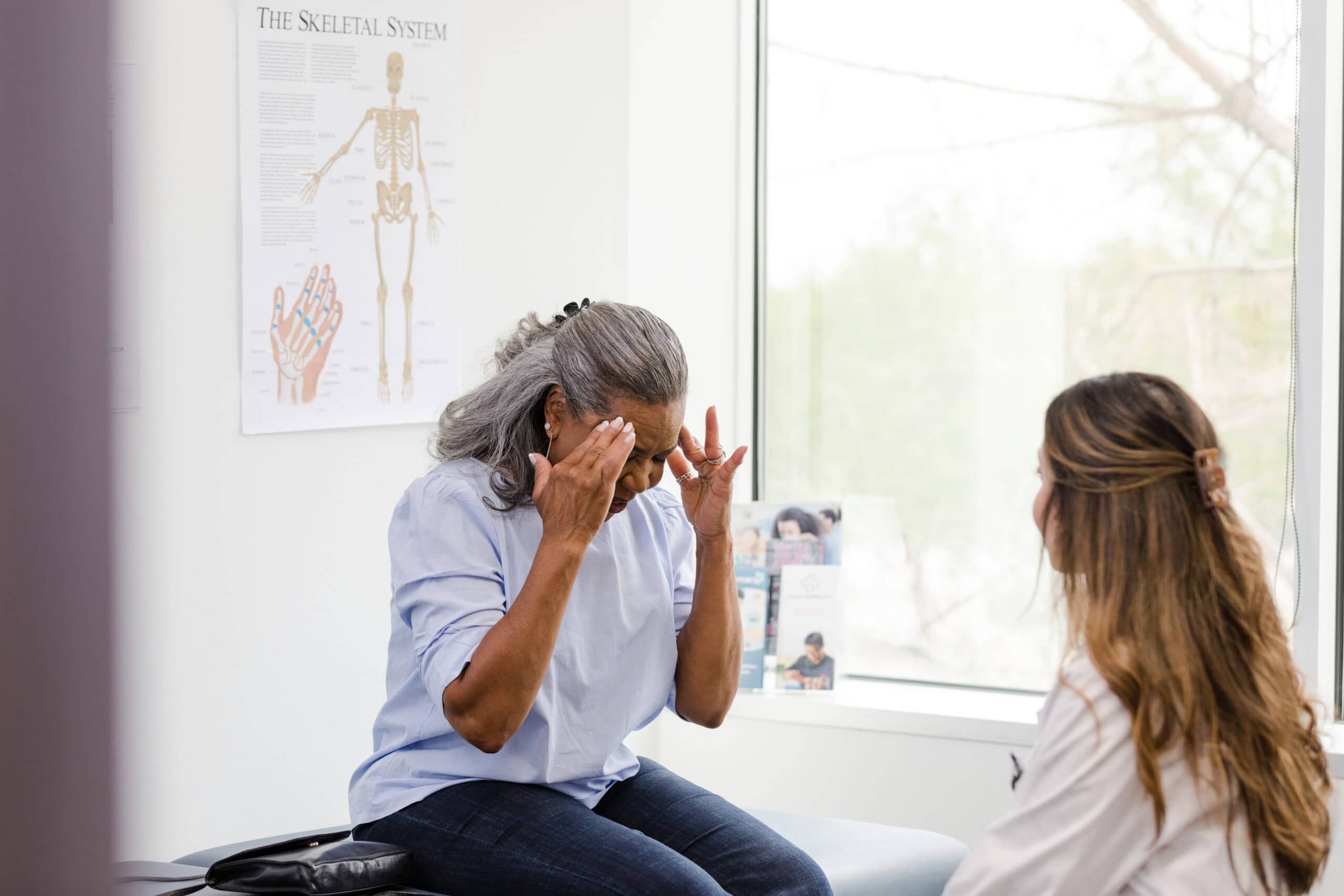 An unrecognizable senior adult woman touches her temples as she explains her headache symptoms to the unrecognizable female doctor.