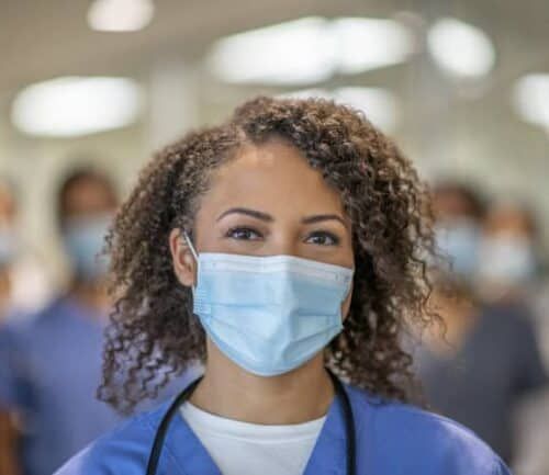 A female doctor is wearing blue medical scrubs and a face mask stands in front of her colleagues at the hospital. She is smiling behind her mask.