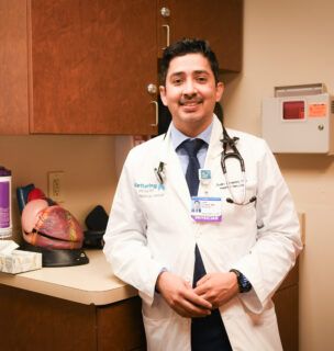 Dr. Juan Linares standing in a white lab coat in a patient room in front of a model of the heart