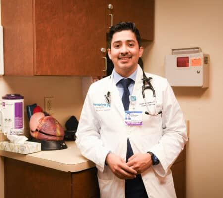 Dr. Juan Linares standing in a white lab coat in a patient room in front of a model of the heart