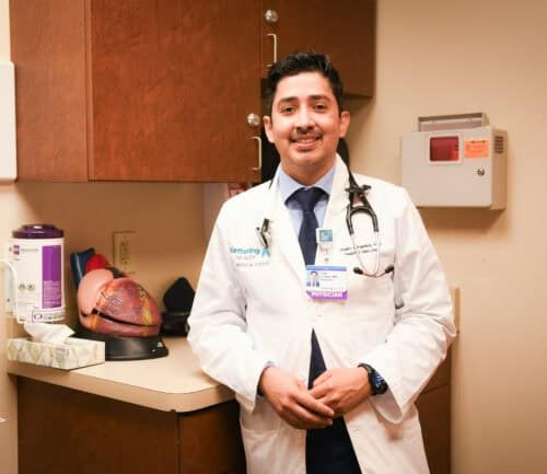 Dr. Juan Linares standing in a white lab coat in a patient room in front of a model of the heart