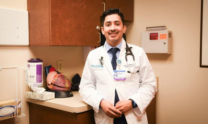 Dr. Juan Linares standing in a white lab coat in a patient room in front of a model of the heart