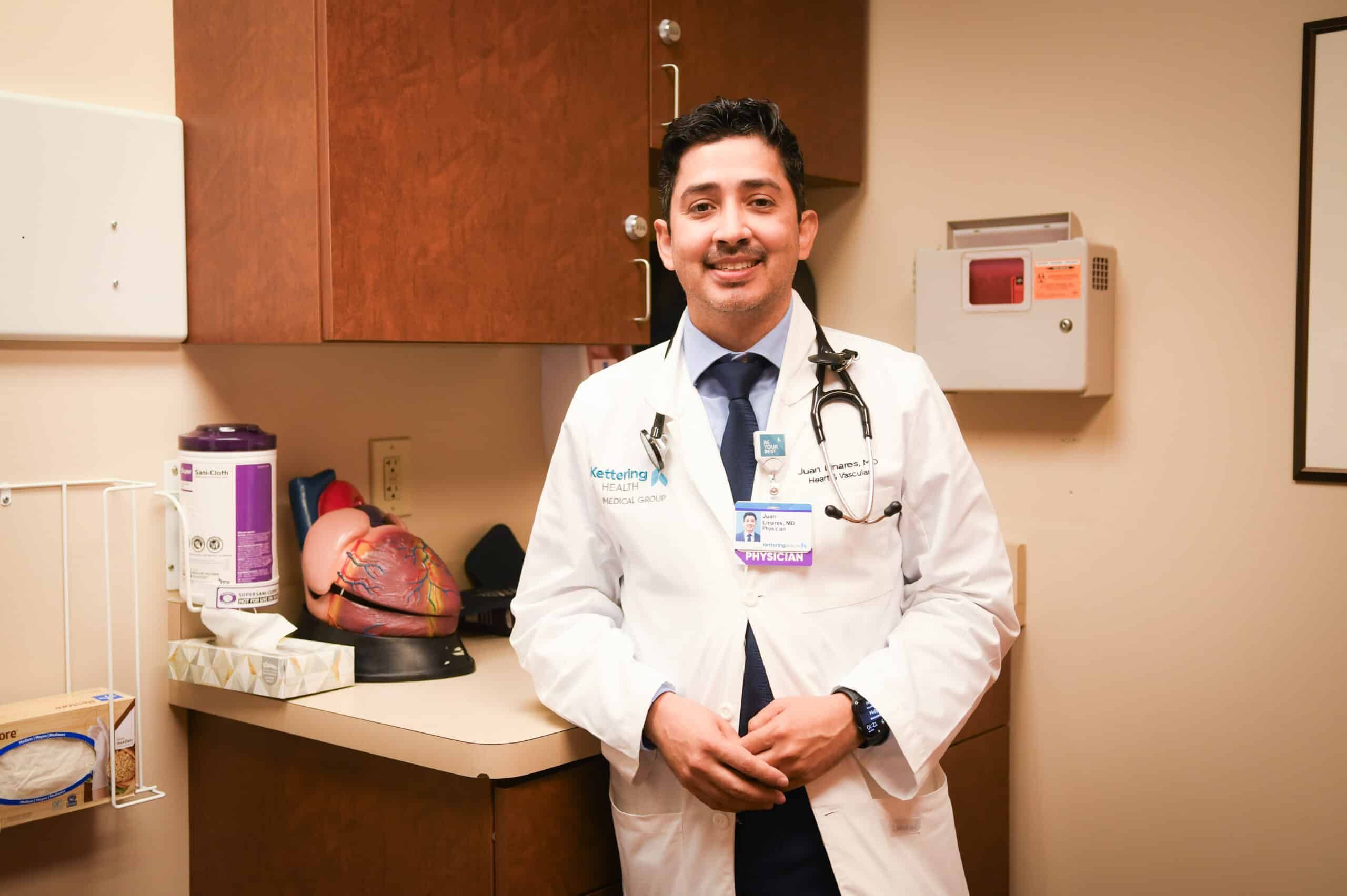 Dr. Juan Linares standing in a white lab coat in a patient room in front of a model of the heart