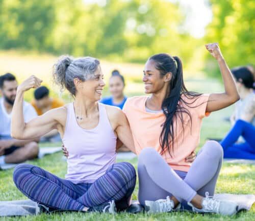 older woman and younger woman sitting outside, flexing their biceps
