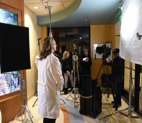 Healthcare professional in white coat being filmed by camera crew in medical facility