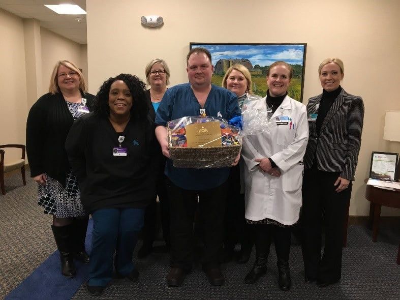 Group of healthcare staff members presenting gift basket to male colleague in scrubs