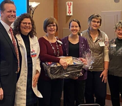 Six healthcare professionals posing together with gift basket near Christmas tree at awards ceremony.