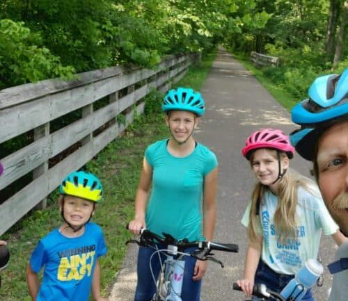Family of five wearing bike helmets posing together on tree-lined bike path