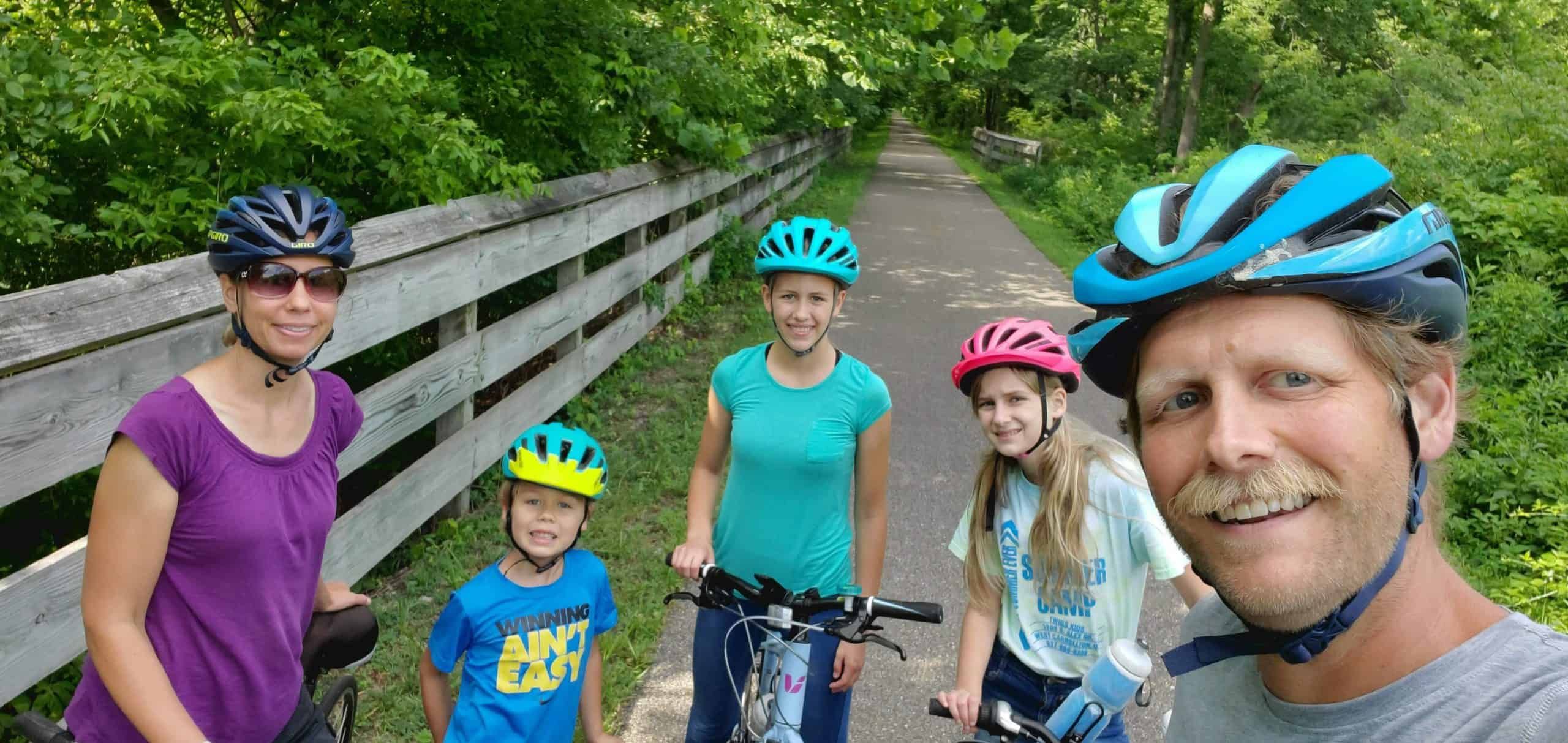 Family of five wearing bike helmets posing together on tree-lined bike path