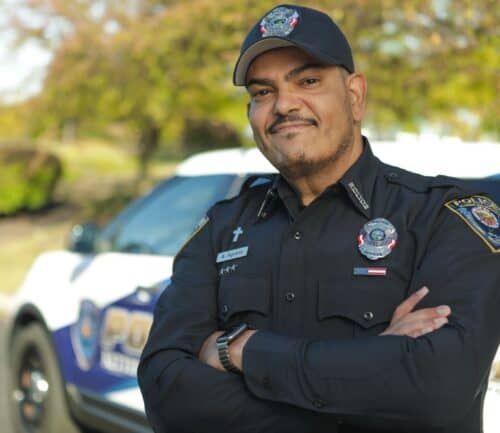 Smiling police officer in uniform with arms crossed standing beside patrol car