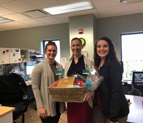 Three women smiling while holding a wicker gift basket in office setting