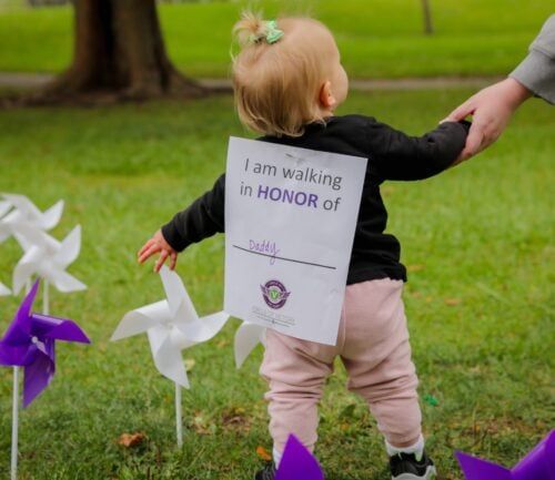 Young child walking with adult holding sign reading "I am walking in HONOR of Daddy"