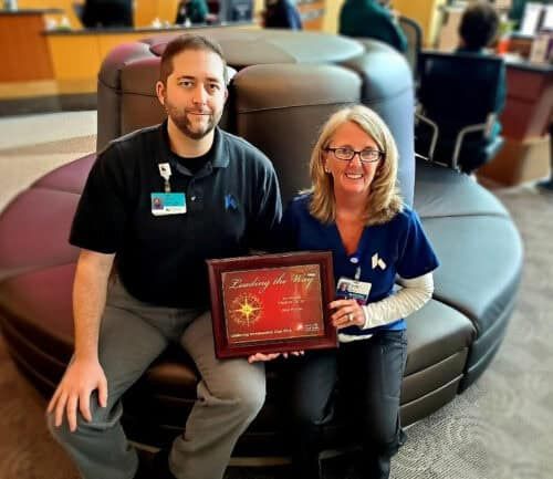 Two healthcare workers sitting together holding a "Leading the Way" recognition plaque
