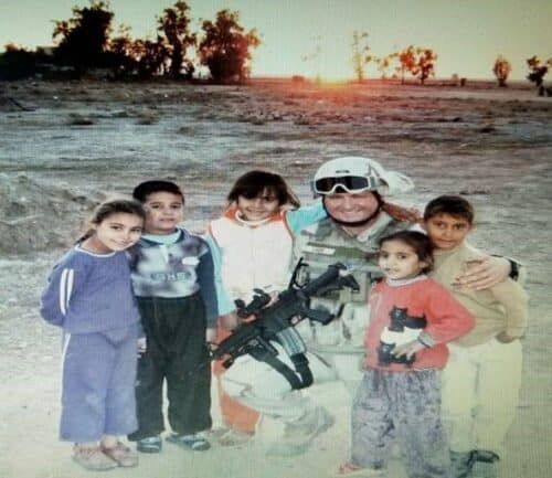 Military service member in combat gear posing with local children in desert setting