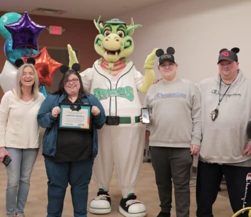 Five people celebrating with Dragons mascot, holding certificate and balloons at healthcare event