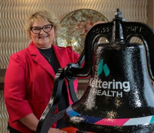 Woman in red blazer stands next to large black Kettering Health bell