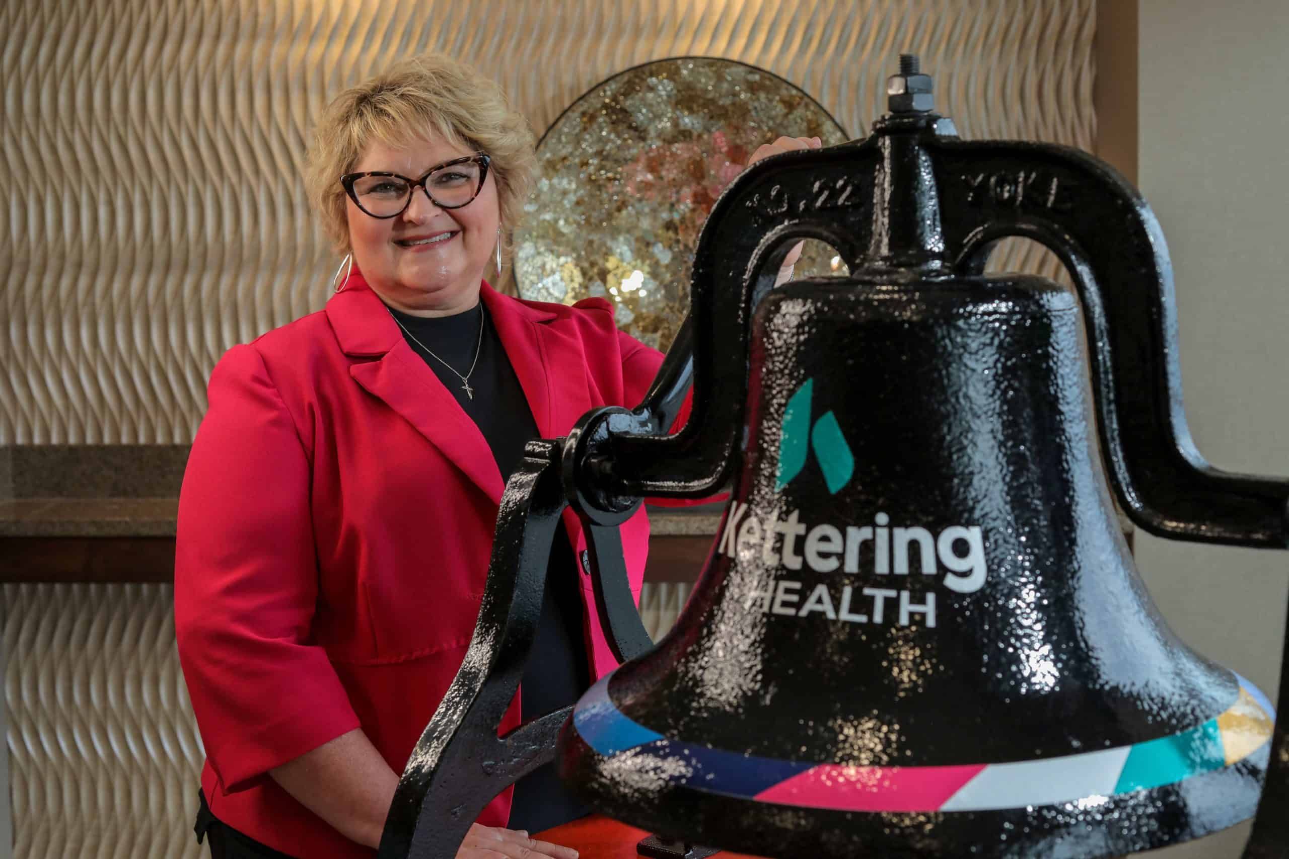 Woman in red blazer stands next to large black Kettering Health bell