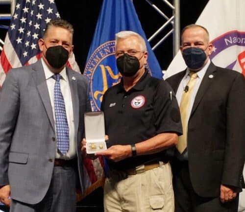 Three masked officials at award ceremony, center person holding recognition box with flags behind
