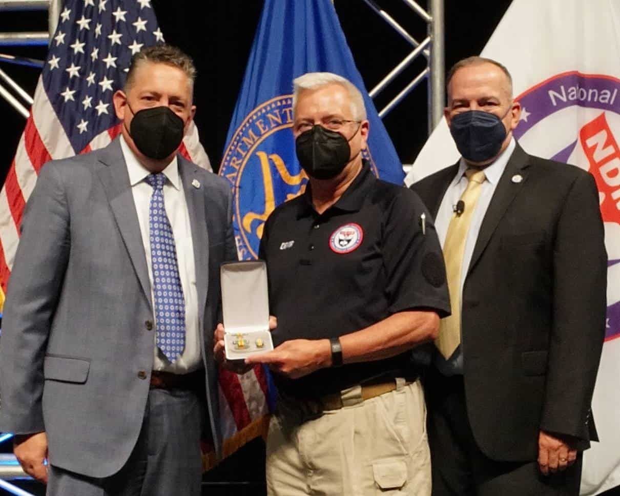 Three masked officials at award ceremony, center person holding recognition box with flags behind