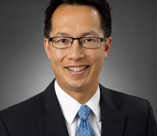 Professional headshot of smiling man in dark suit with blue patterned tie and glasses