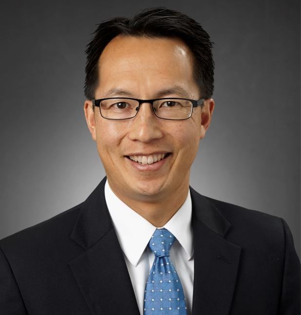 Professional headshot of smiling man in dark suit with blue patterned tie and glasses