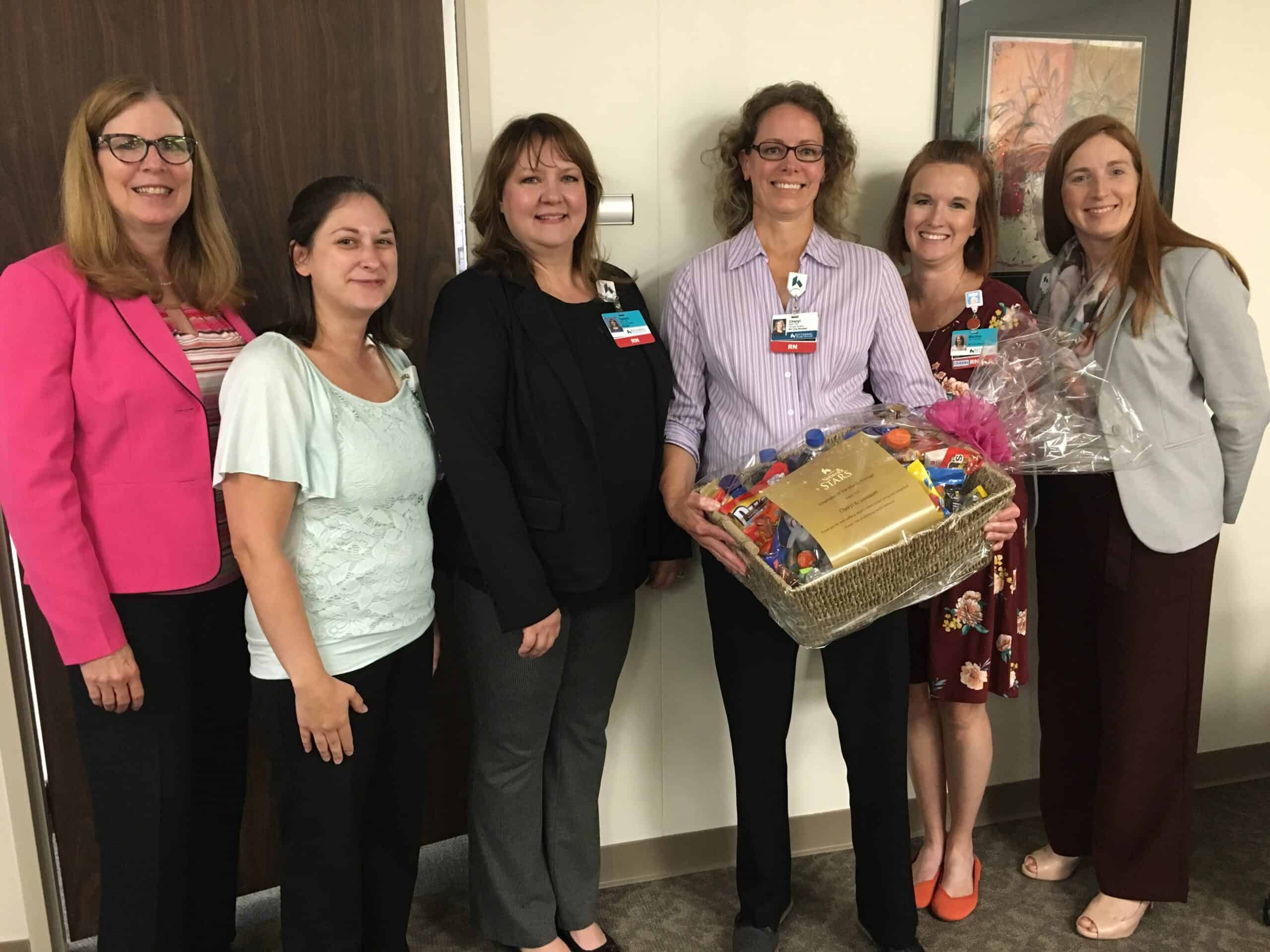 Six healthcare professionals posing together, one holding a large gift basket with chocolates and treats.