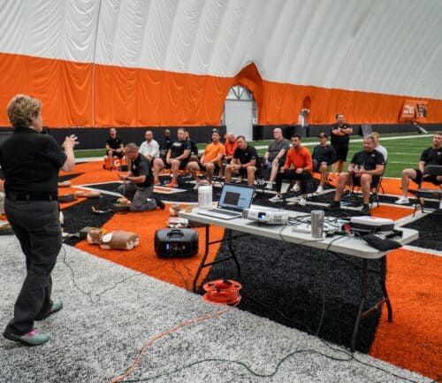 Medical training session with CPR mannequins on football field, instructor presenting to seated group