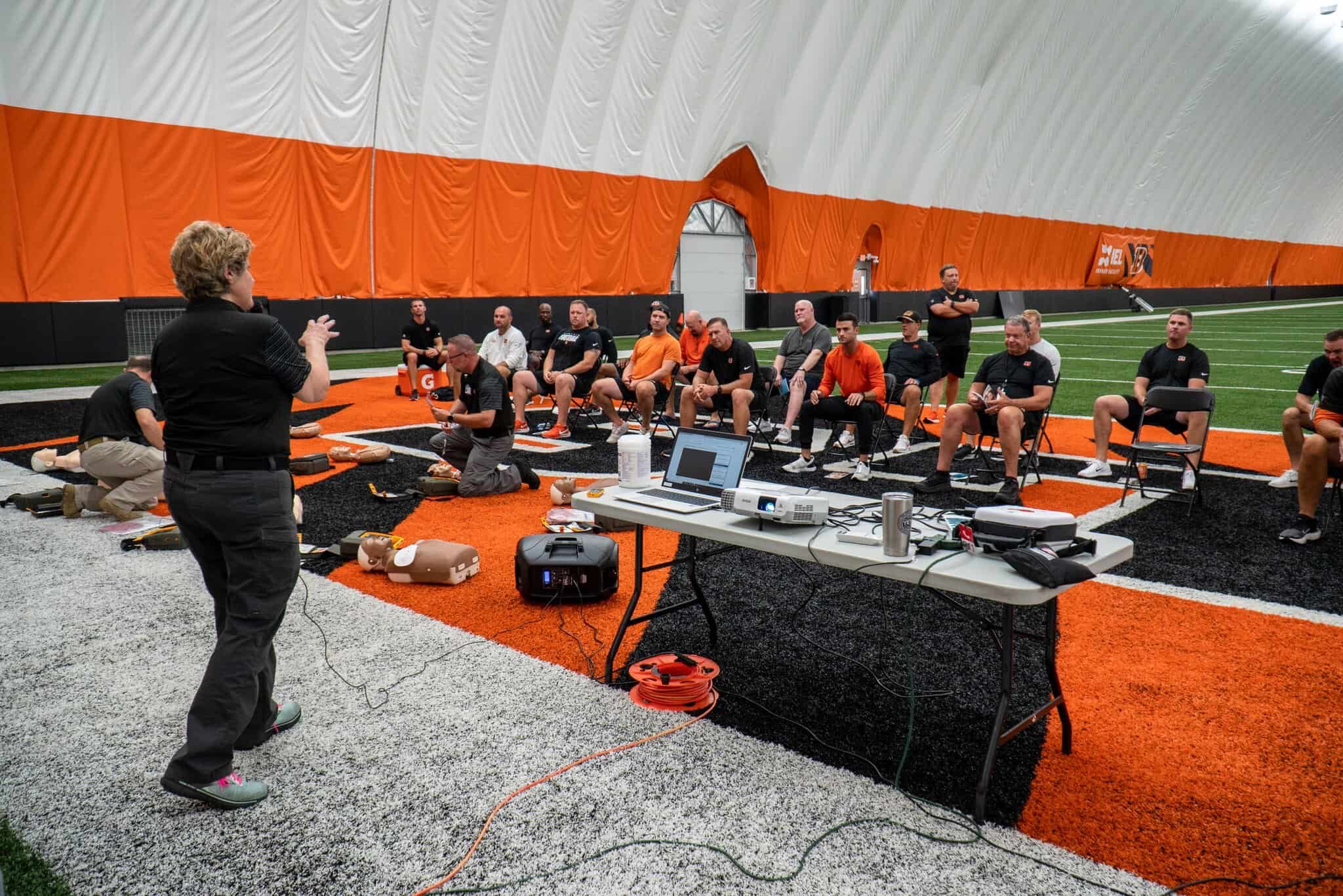 Medical training session with CPR mannequins on football field, instructor presenting to seated group