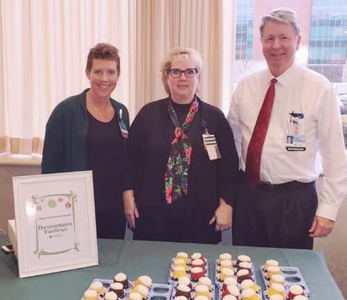 Three healthcare staff members standing behind table with cupcakes and documentation excellence award sign
