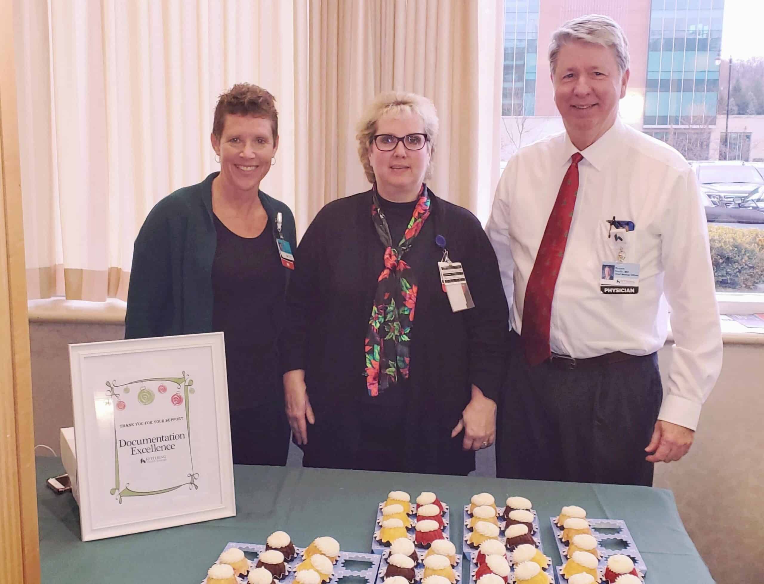 Three healthcare staff members standing behind table with cupcakes and documentation excellence award sign