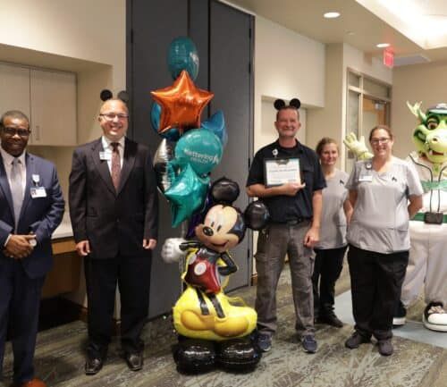 Healthcare staff and community members celebrating with Mickey Mouse balloons and mascot