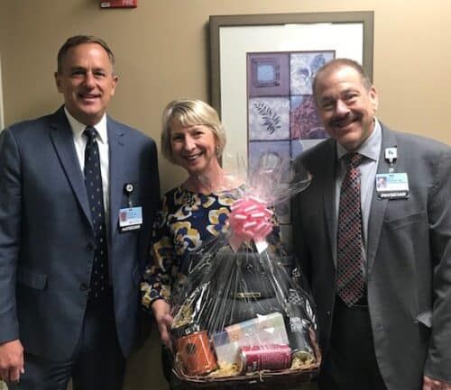 Two men in suits and woman holding gift basket pose together in medical facility