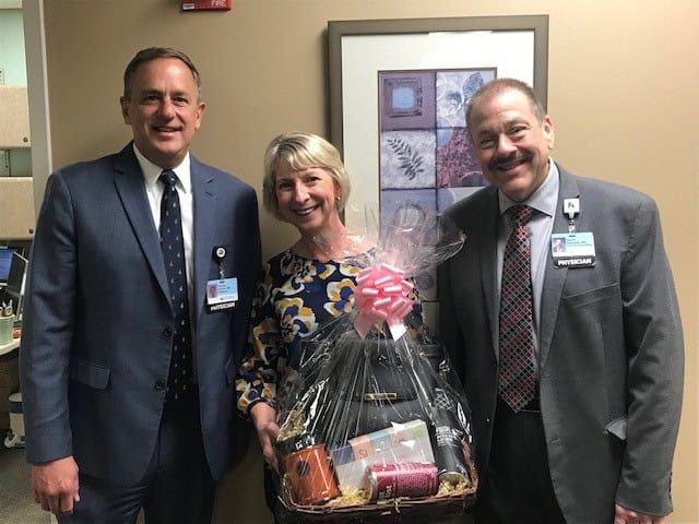 Two men in suits and woman holding gift basket pose together in medical facility