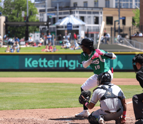 Baseball player in green jersey swinging bat at Kettering Health sponsored stadium