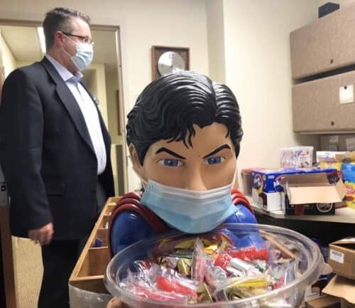 Man in suit observing Superman figure wearing mask while holding bowl of candy