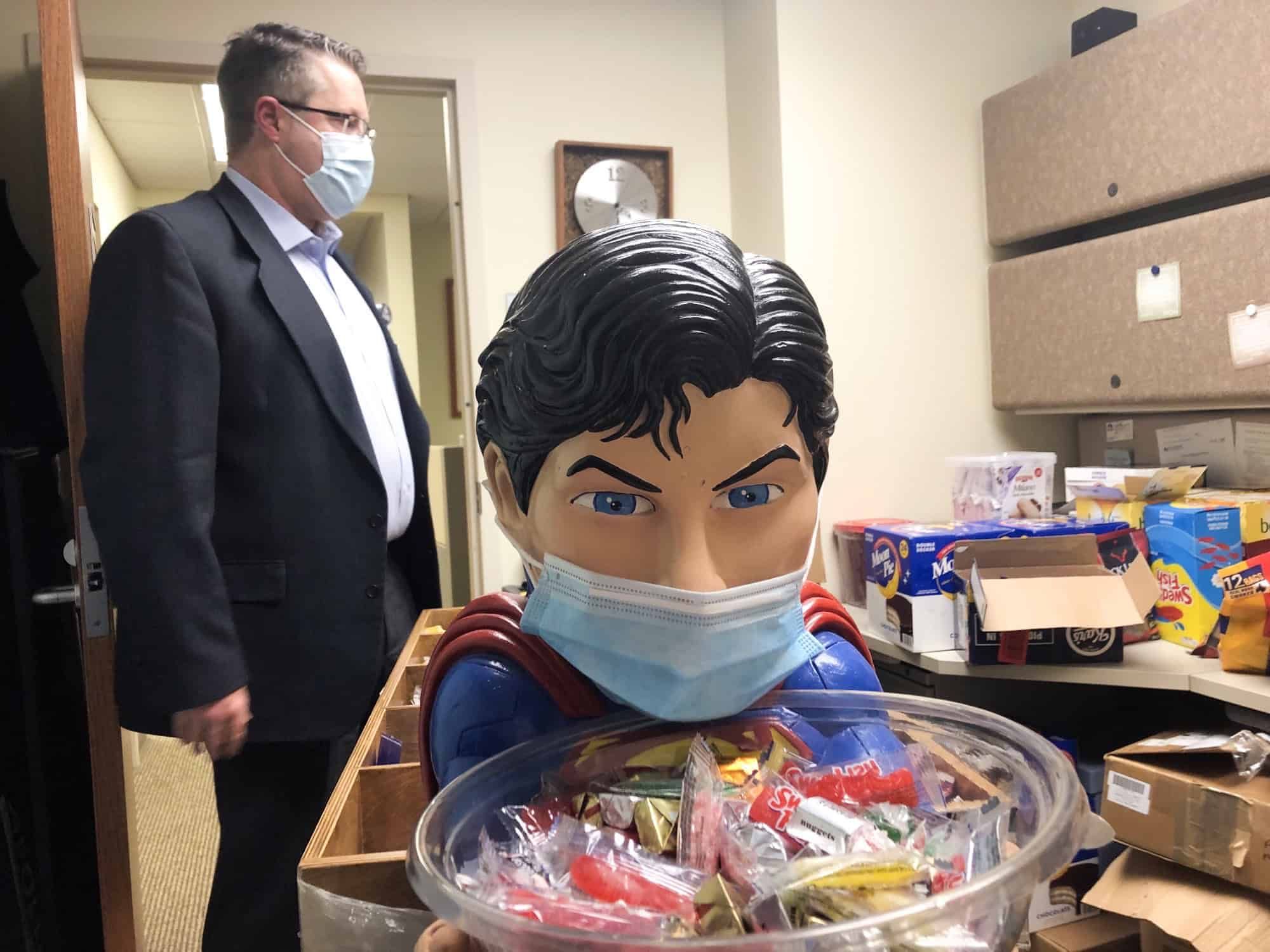 Man in suit observing Superman figure wearing mask while holding bowl of candy