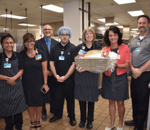 Seven healthcare staff members posing together in hospital kitchen, one holding gift basket