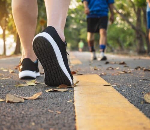 Runner's feet in black sneakers on yellow-lined asphalt path with fallen leaves