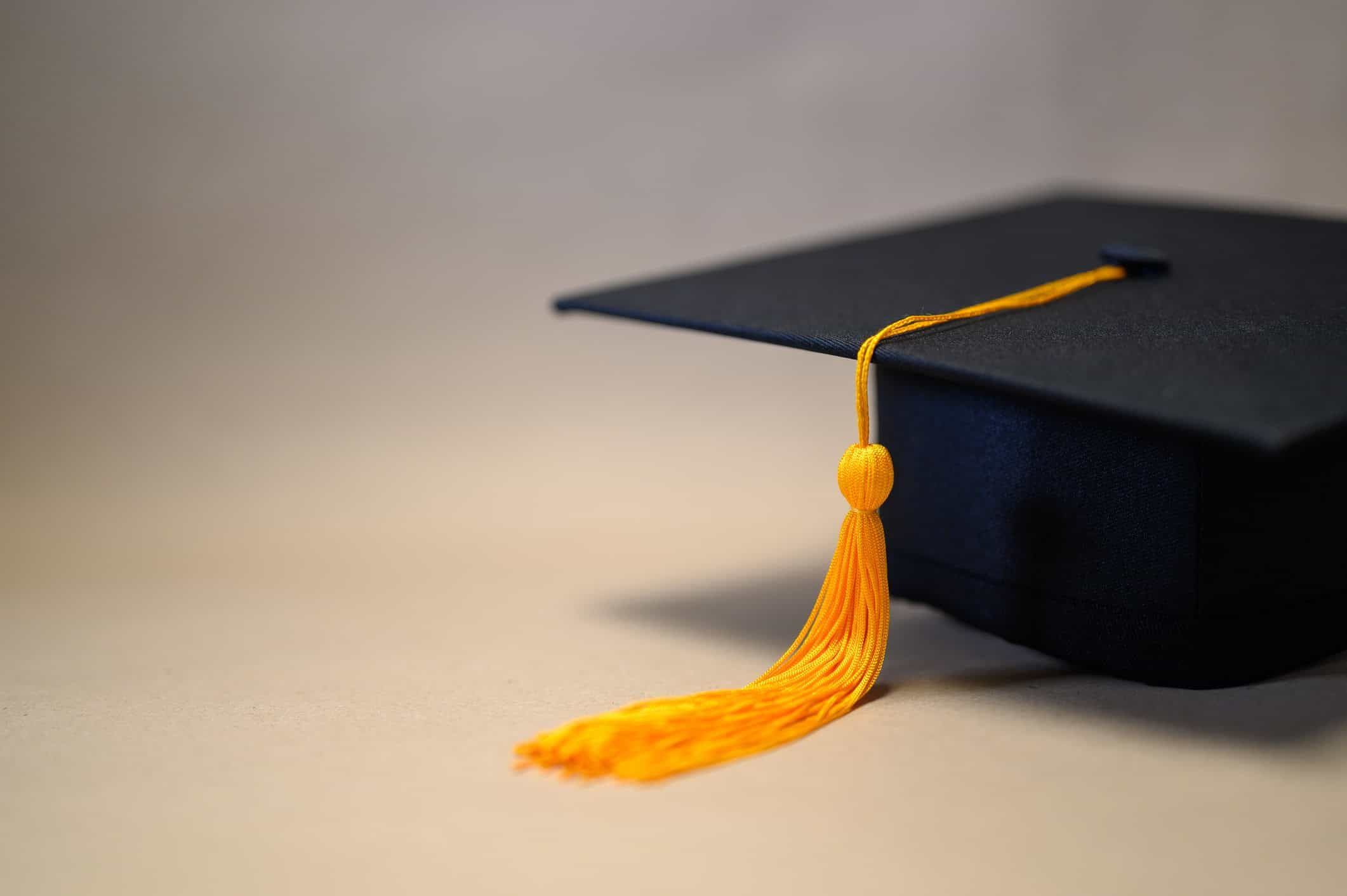 Black graduation cap with gold tassel on neutral background