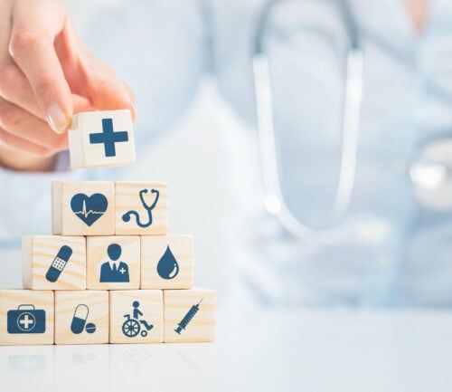 Doctor's hand stacking wooden blocks with medical icons including cross, stethoscope, and syringe