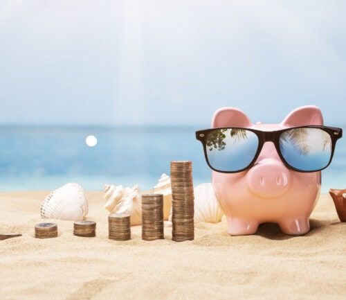 Pink piggy bank wearing sunglasses on beach sand beside stacked coins and seashells