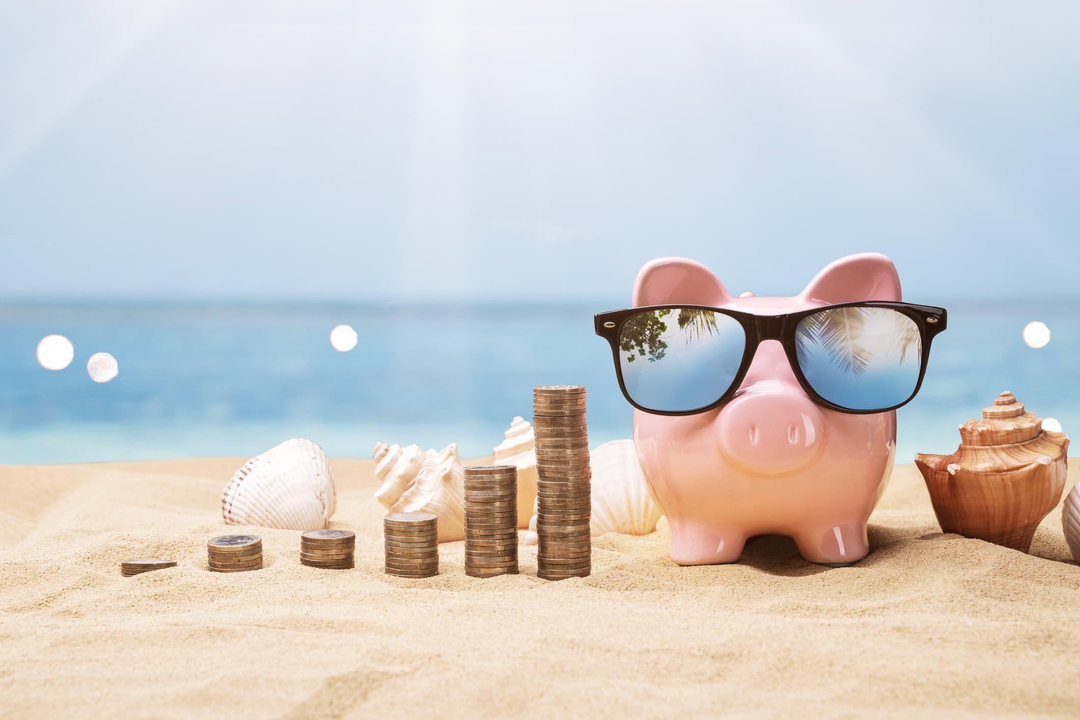 Pink piggy bank wearing sunglasses on beach sand beside stacked coins and seashells