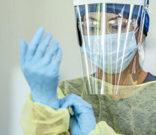 Hispanic female healthcare worker in PPE raising hand behind protective barrier