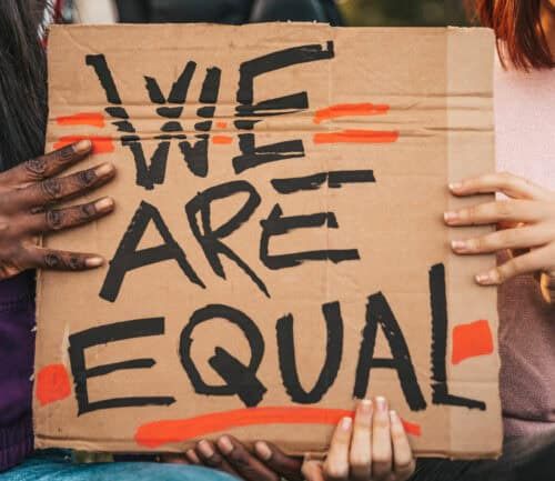 Multiple hands holding cardboard sign reading "WE ARE EQUAL" with orange highlighting